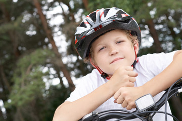 Small Caucasian boy cyclist in protective helmet put his head on the handlebar of the bike posing for the camera. a Boy rests during cycling in park on summer day. Weekend activity.