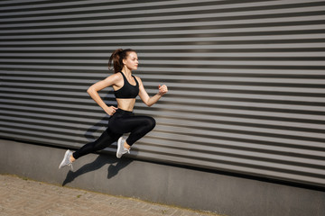 Young woman with fit body jumping and running against grey background. Female model in sportswear exercising outdoors