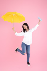 Asian teenager girl holding yellow umbrella during cold rainy day,drinks hot coffee to warm herself,  jumping on pink background.  Autumn concept