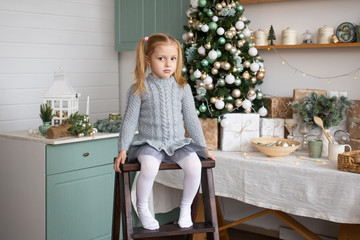 girl is sitting near decorated Christmas tree in kitchen indoors.