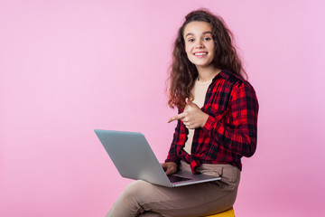 Naklejka premium Portrait of excited teenage girl with curly brunette hair in plaid shirt pointing at laptop and smiling happily, satisfied with computer software. indoor studio shot isolated on pink background
