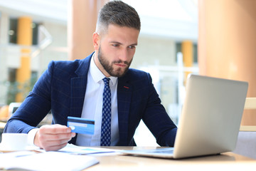 Smiling man sitting in office and pays by credit card with his laptop.