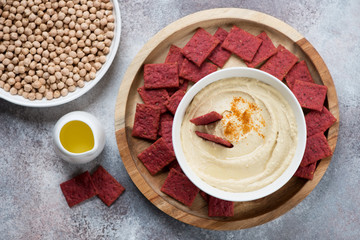 Round wooden tray with a bowl of hummus and beetroot crackers, flatlay on a beige stone background