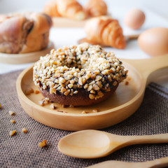 Homemade food fresh bakery, Closeup delicious chocolate donut almond peanut topping served on wooden dish, spoon, fork on napery and white kitchen table with bread bun croissant and eggs background