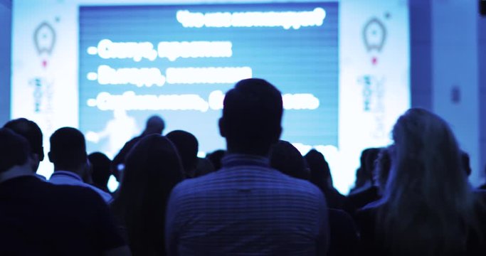 Business conference, training and forum. Viewers looking at presentation on screen and listening man speaker talking from stage. Public speech concept. Educational program for entrepreneurs.