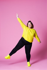 Cheerful plus size model in casual clothes, fat woman in black jeans and yellow jumper on pink...
