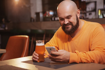 Cheerful bearded man using his smart phone while drinking beer at the pub