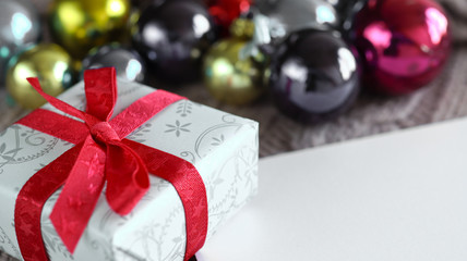Christmas gift with red ribbon and bow, on the blurred background with colorful christmas balls and copy space