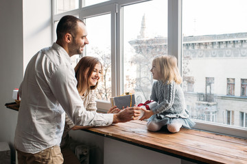 Parents present their little daughter gift box at home on window. Merry Christmas and Happy New Year!