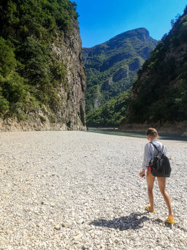 Young woman with a backpack crosses the small river, flowing between tall mountain peaks.A girl walks along the shore between the mountains, Albania Shkoder, Komani river.