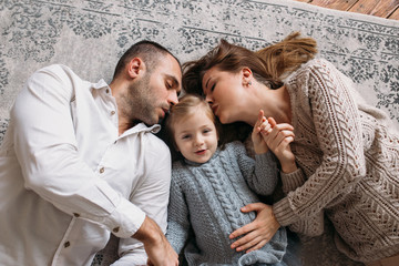 Parents kissing their little daughter lying on the floor at home.