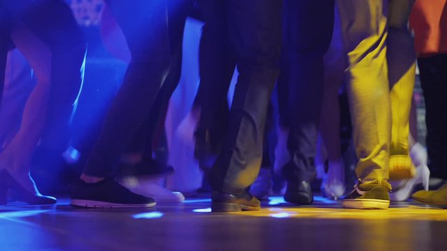 Many female and male legs energetically dance at a disco in the nightclub in the rays of lamps.