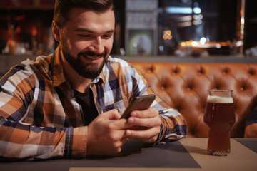 Charming bearded man texting on his smart phone at the beer pub. Attractive man using smart phone while having glass of beer