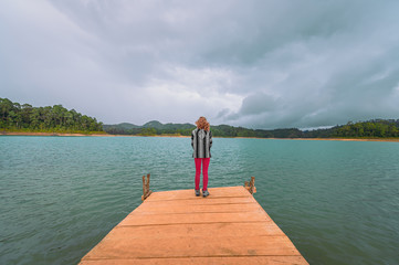 Young blonde woman on a pier watching a turquoise lake and background mountains with forest