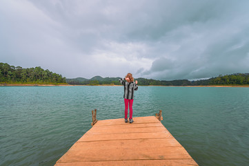 Fototapeta na wymiar Young blonde woman on a pier watching a turquoise lake and background mountains with forest