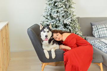 Pretty young girl in red dress siiting on bed and hugs her husky dog over christmas tree