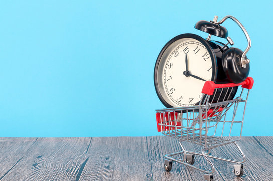 Shopping cart and classic alarm clock on blue background. Sale time buy mall market shop consumer concept. Selective focus.