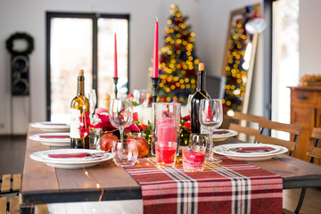 Table served for Christmas dinner with decorations, candles, fruits, glasses and lanterns. Bright living room decorated with lights and Christmas tree. Natural light shot.
