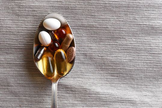 Close-up tablespoon filled with various dietary supplements, tablets and vitamins on a gray fabric background