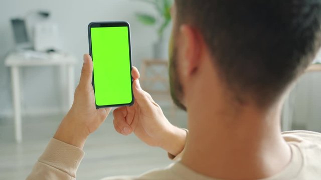 Slow motion of bearded guy brunet is using smartphone with green screen in house holding gadget in hands being in apartment alone. People and devices concept.