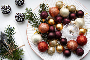 Christmas colored decorations lie on a white plate along with the branches of the Christmas tree. A white napkin and candles stand nearby.