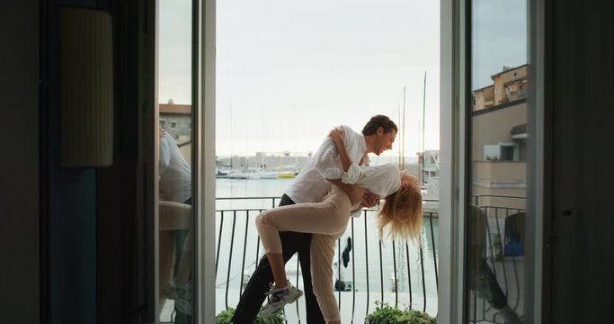 A cheerful young couple is enjoying time together on a terrace of a hotel room during their romantic vacation with panoramic view on a sea with yachts in a sunny day.