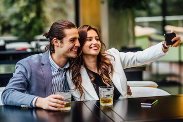 Lovely concept, pretty young and cheerful couple with drinks on table spend time at modern restaurant and make a selfie.