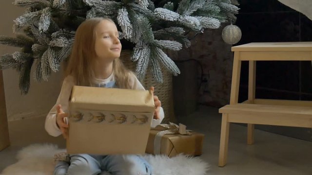 Cute blond girl has recieved a gift box wrapped in craft paper sitting near christmas tree, happy childhood and xmas celebration concepts
