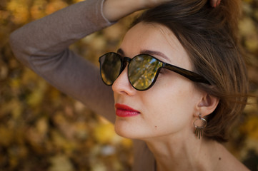 Young and beautiful girl with short hair and sunglasses with mirror surface is posing against the trees with yellow leaves background spending time in the autumn park