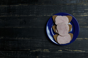 Sandwich of dark bread and sausage slices on blue plate standing on black wooden background. Tasty unhealthy food. Diet, nutrition, eating habits concept. Place for text