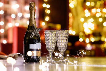 Bottle of champagne and two glass goblets stand on the backdrop of warm white lights on Christmas night. Concept for a romantic new year celebration. Holiday Promotions and Gifts