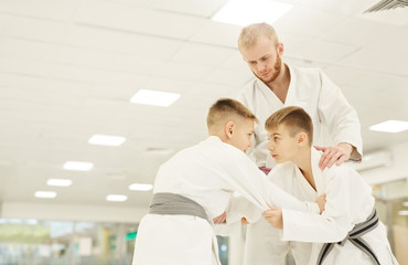Fototapeta premium Two boys in kimono practicing technique with their coach standing near by and teaching them during training in gym