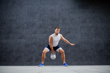 Strong muscular handsome Caucasian man in shorts and t-shirt standing outdoors and swinging kettle bell. In background is gray wall.