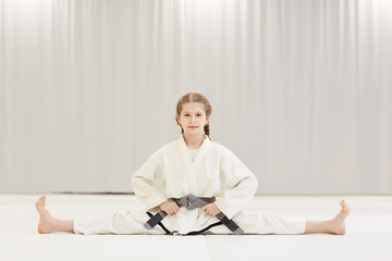 Portrait of little girl in white kimono sitting on the twine and doing stretching exercises during training in karate