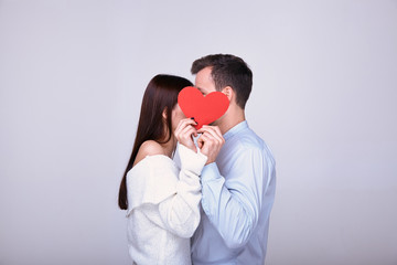 Beautiful couple of lovers stands on a white background and covers their faces with a red heart