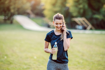Fototapeta premium Beautiful fit smiling caucasian woman in sportswear and with ponytail standing in nature, talking on the phone and holding bottle with refreshment. Fitness in nature concept.