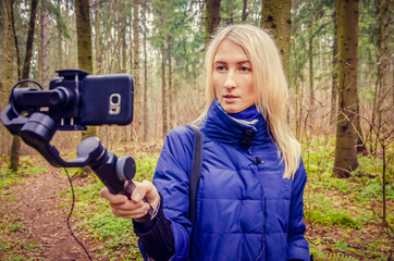 Young blonde girl blogger's shooting a video on a mobile phone and gimbal in the woods. concept of blogging, information, modern technologies.