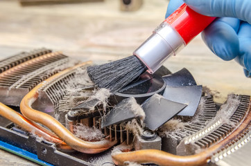 man's hand in blue gloves with a brush cleans the cooler of the video card close-up. Dust on the CPU cooler of the computer's graphics card close-up on a wooden table.