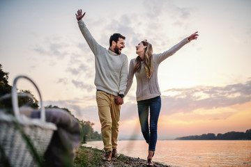 Playful caucasian couple holding hands and walking on coast next to river. Both are dresses in white sweaters. In foreground in picnic basket.