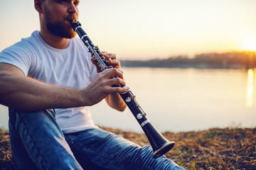 Young handsome bearded caucasian blond man sitting on cliff and playing clarinet. In background is...