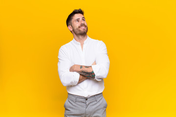 young handosme man feeling happy, proud and hopeful, wondering or thinking, looking up to copy space with crossed arms against flat color wall