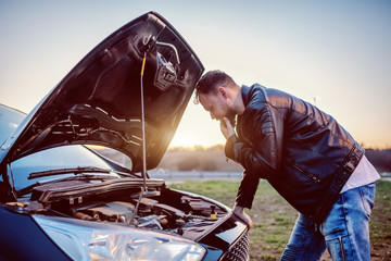 Young serious handsome caucasian bearded blond man in leather jacket looking engine of his car...