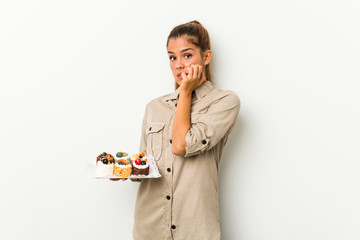 Young caucasian woman holding sweet cakes biting fingernails, nervous and very anxious.