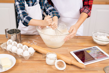 Little boy whisking flour with raw eggs in bowl by table while helping mom