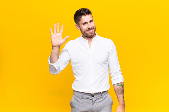 young handosme man smiling happily and cheerfully, waving hand, welcoming and greeting you, or saying goodbye against flat color wall