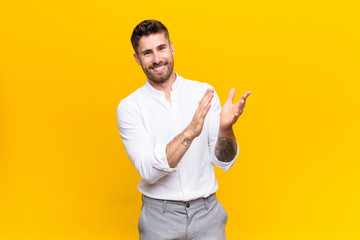 young handosme man feeling happy and successful, smiling and clapping hands, saying congratulations with an applause against flat color wall