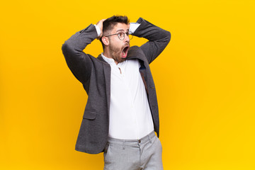 young handosme man with open mouth, looking horrified and shocked because of a terrible mistake, raising hands to head against flat color wall