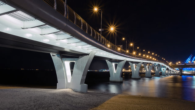 a bridge over the Neva river in St. Petersburg, a pedestrian bridge intertwined with the ZSD toll highway and a view of a large beautiful stadium in the background.