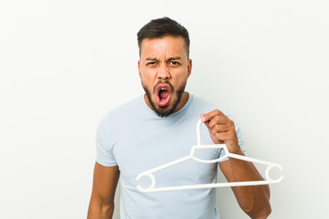 Young south asian man holding a hanger screaming very angry and aggressive.