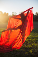 The slender gymnast in black dress in the middle of the field choreographs a large piece of red chiffon in the sunset light.